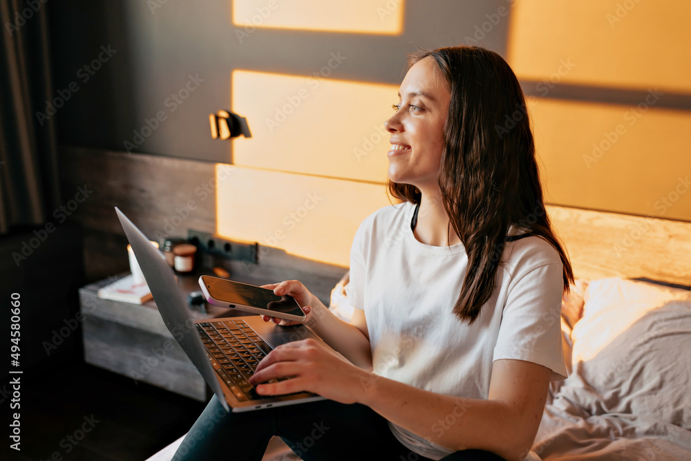Attractive smiling lovely girl with dark wavy hairstyle is working with laptop in the bed and looking at window in modeming sunshine. Caucasian young woman Is scrolling smartphone and holding laptop