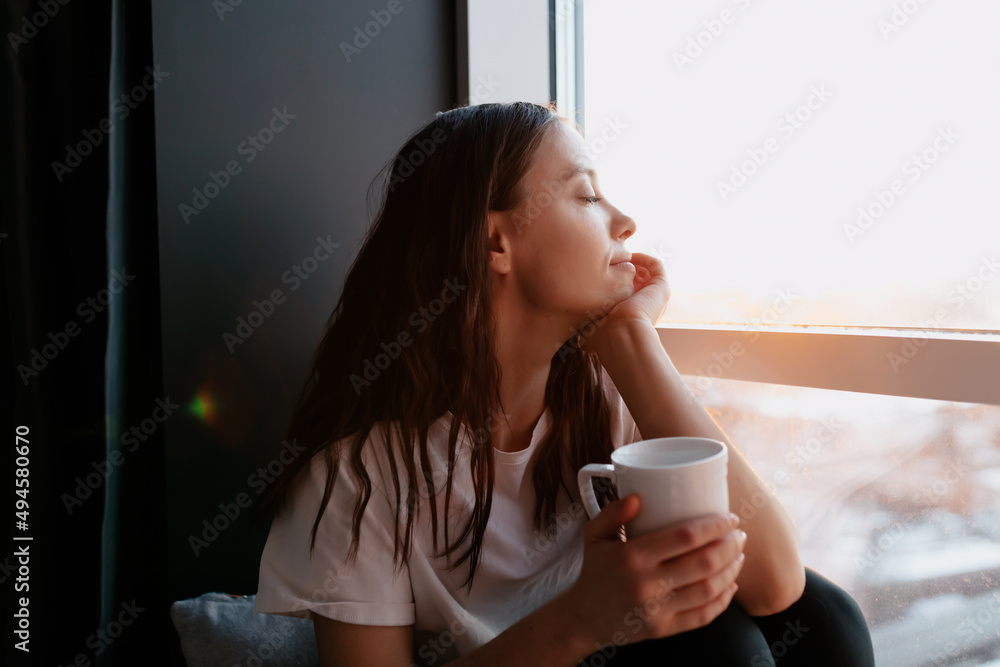 Close up portrait of romantic lovely woman with dark hair with wavy hair is closed eyes and smiling while drinking morning coffee and sitting near the window in morning sunshine