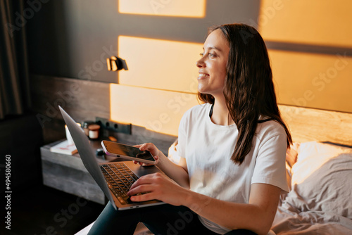 Obraz na płótnie Attractive smiling lovely girl with dark wavy hairstyle is working with laptop in the bed and looking at window in modeming sunshine. Caucasian young woman Is scrolling smartphone and holding laptop w salonie