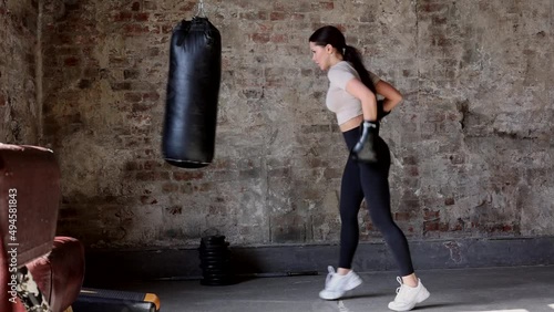 beautiful middle-aged thin sporty woman boxing and bitting punching bag. strong muscular young girl practicing box in black boxing gloves in gym. Fit slim woman in gris gym in black pants photo
