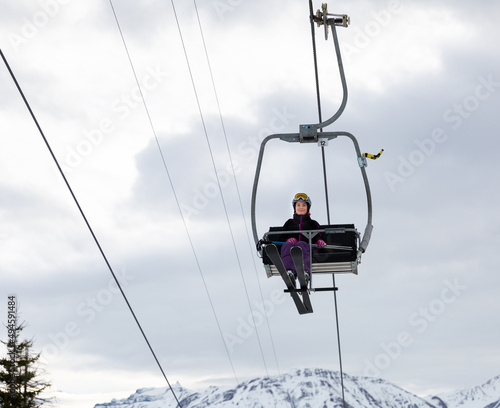 Smiling woman skier in full ski equipment riding on chairlift, lifting to top of mountain to pistes on background of snowy peaks on cloudy winter day..