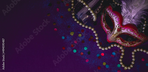 Masquerade mask, white pearl beads, confetti and an empty champagne glass on a dark lilac.