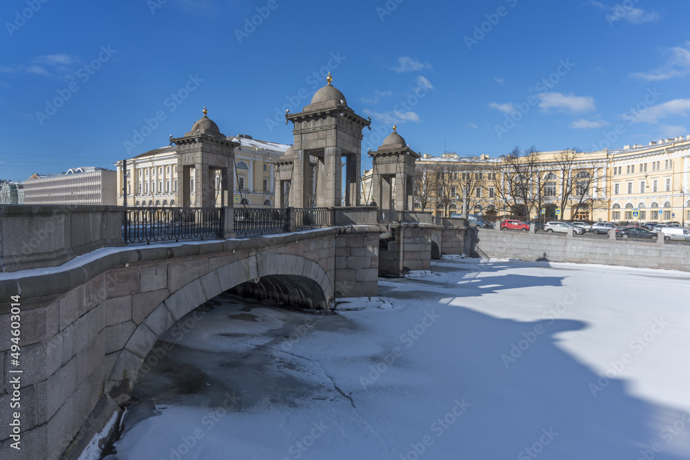 View of the ancient stone Lomonosov bridge with frozen river in St. Petersburg, Russia in spring. The bridge is decorated with 4 towers with chains and domes. Blue-white sky and old city architecture