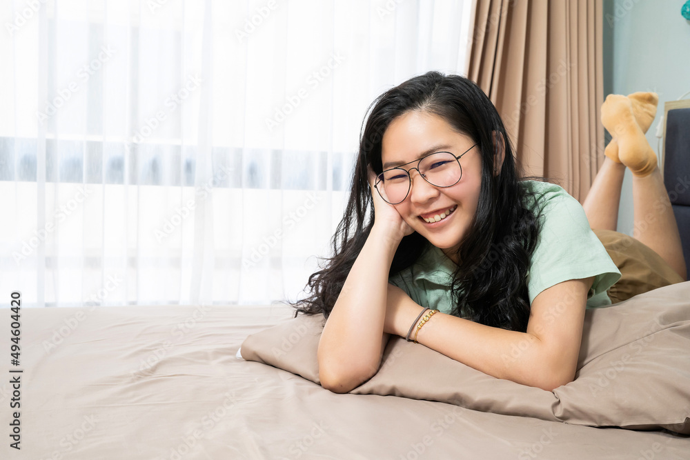 Beautiful glasses Asian woman is lying on her stomach and looks at camera on her bed in the bedroom with a pastel green - brown color theme. Beautiful glasses Asian woman is lying on her stomach and looks at camera on her bed in the bedroom with a pastel green - brown color theme.