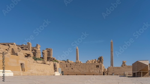Paved square, dilapidated walls of the ancient Karnak temple in Luxor. Two tall obelisks against a clear blue sky. Copy space. Egypt