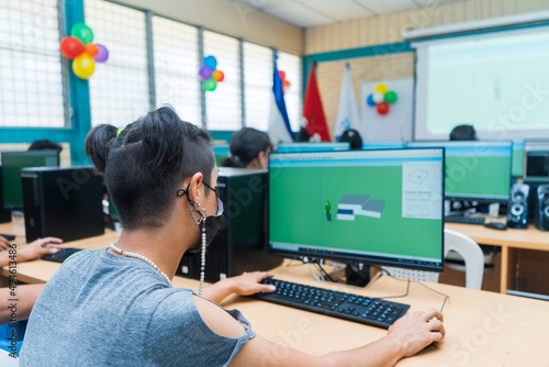 Young Central American student woman with light skin and a modern hairstyle seen from the back receives technical education and uses a desktop computer to do her homework in a laboratory.