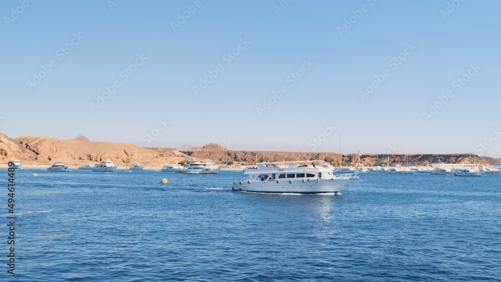 Large white yacht sailing across blue sea on sunny day, against marina backdrop with anchored yachts. Luxury motor yacht side view emerging from ship dock. Egypt Red Sea Cruise. Holiday yacht vacation