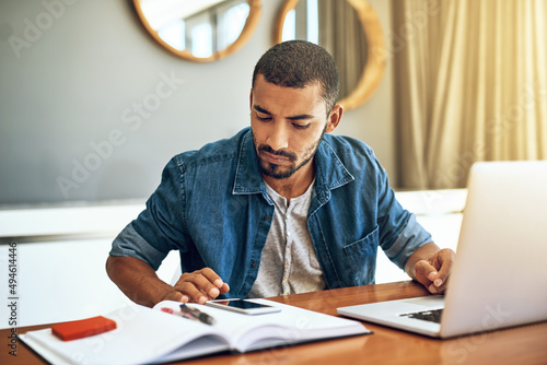 Crunching the numbers. Shot of a focused young man working on his laptop and checking his phone while being seated at a table at home.
