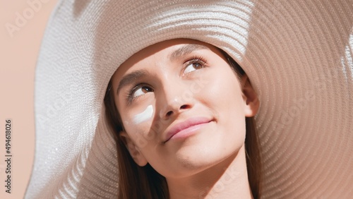 Young pretty brunette European woman in a big white hat smears sunscreen on her cheek and enjoys the sun smiling for the camera against beige background | Sunblock concept