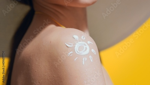 Young dark-haired woman in a big white hat and yellow swimsuit applies sunscreen on her shoulder against yellow background | Sunscreen application | Sun shape