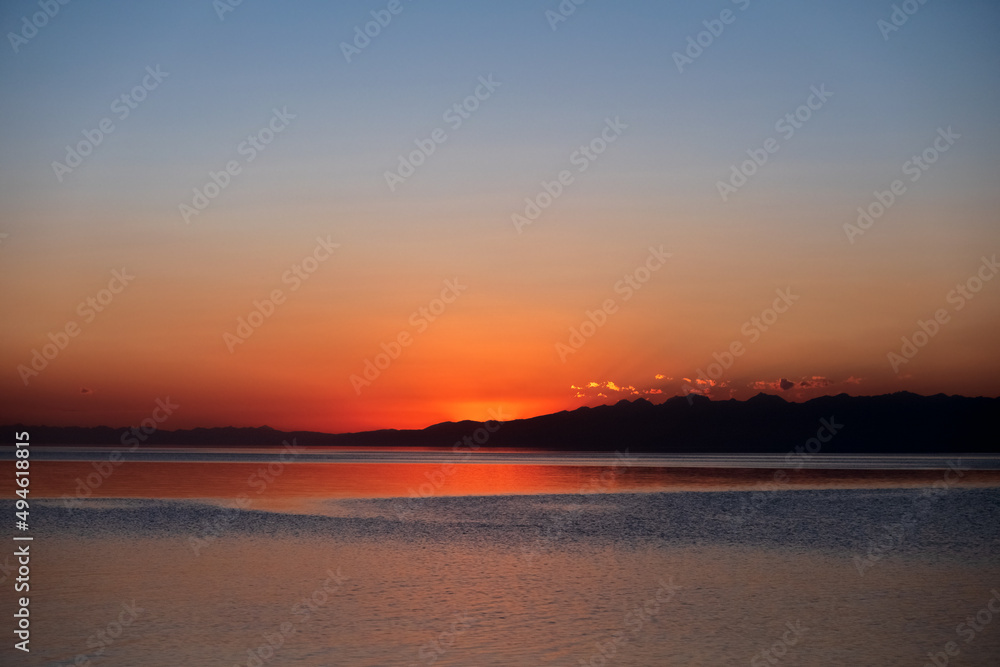 Beautiful sunset on mountain lake with mountain ridge silhouette. Colorful sunrise with orange sky on Issyk-kol lake. Travel, tourism in Kyrgyzstan concept.