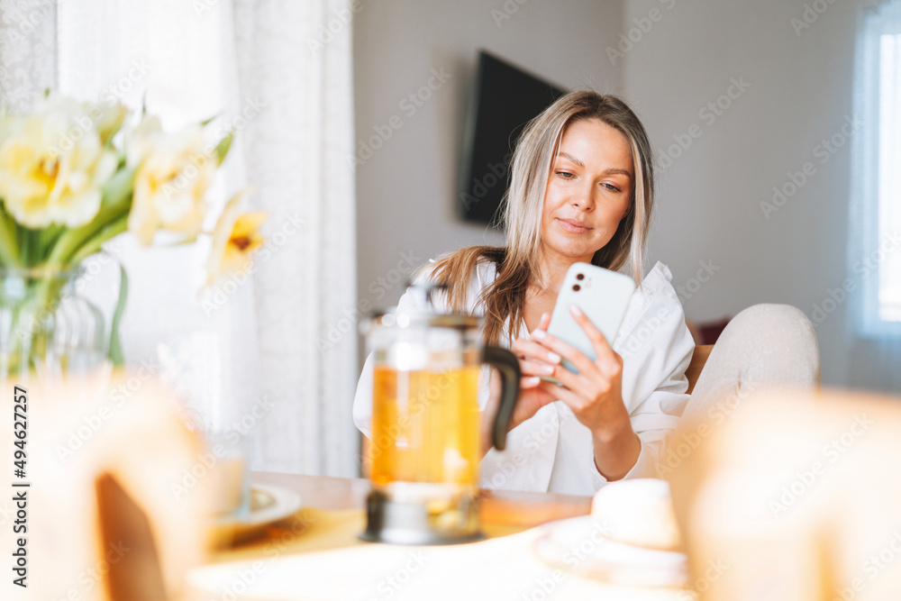 Young beautiful happy woman with blonde long hair in white shirt drinking tea using mobile phone at sunny living room at the home Young beautiful happy woman with blonde long hair in white shirt drinking tea using mobile phone at sunny living room at the home
