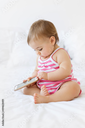 chubby baby girl sitting on a white bed holding smartphone pressing button with finger photo