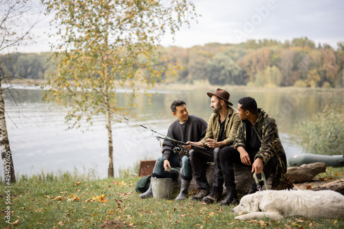 Multiracial male friends resting and talking in nature. Men after fishing on river or lake coast. Concept of leisure and weekend in nature. Idea of friendship and spending time together with dog