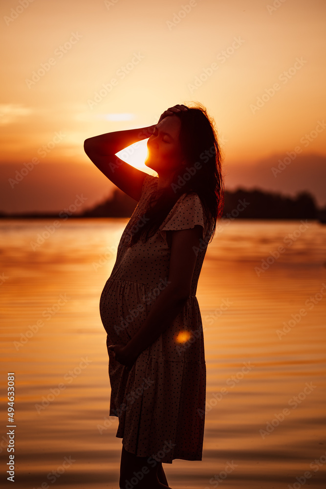Fototapeta premium Silhouette of pregnant woman. Happy young woman in rays of setting sun stands in water on seashore and touches her hair. 