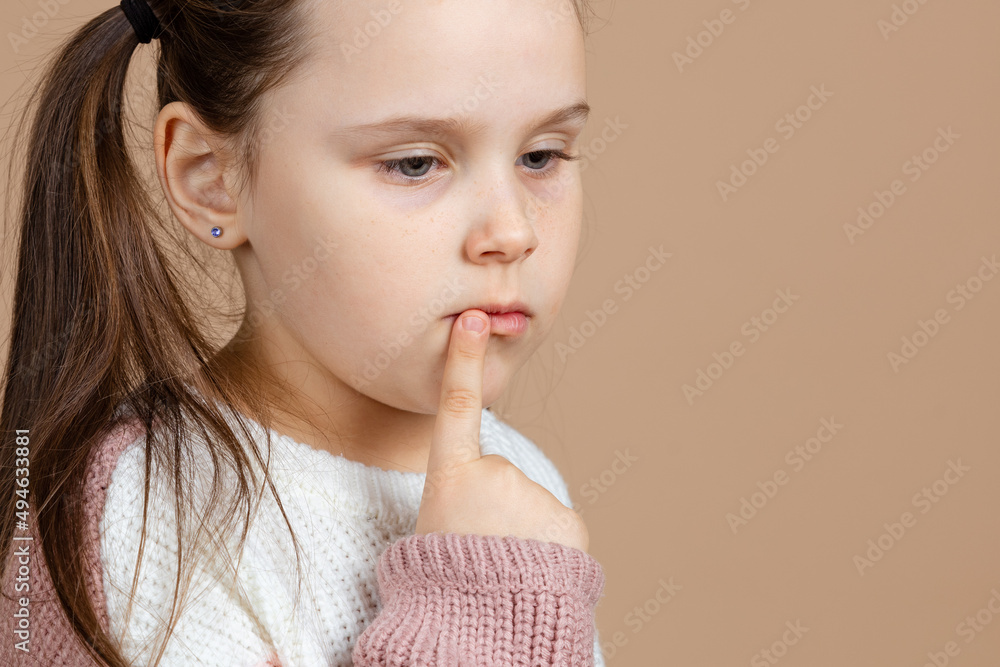Portrait of young cute sad pensive blue-eyed girl with long dark hair in white and pink sweater standing with index finger pointed at mouth, expressing melancholy, gloomy feeling on brown background Portrait of young cute sad pensive blue-eyed girl with long dark hair in white and pink sweater standing with index finger pointed at mouth, expressing melancholy, gloomy feeling on brown background