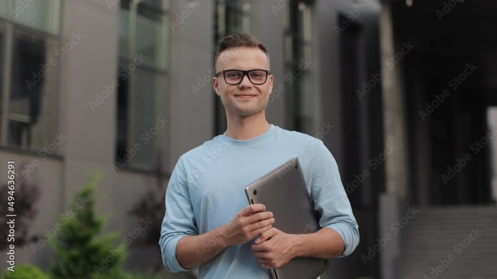 A businessman with a laptop in his hands is standing and looking at the camera. He is wearing glasses and a sweater. He is smiling. He is standing near the business center. 4k A businessman with a laptop in his hands is standing and looking at the camera. He is wearing glasses and a sweater. He is smiling. He is standing near the business center. 4k