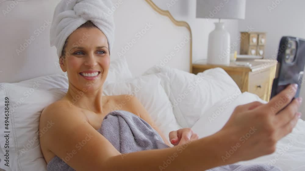 Mature woman lying on bed at home posing for selfie on mobile phone and laughing - shot in slow motion Mature woman lying on bed at home posing for selfie on mobile phone and laughing - shot in slow motion