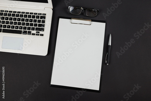 Top view photo of black clipboard folder with white paper sheet, pen, glasses and laptop on isolated black background