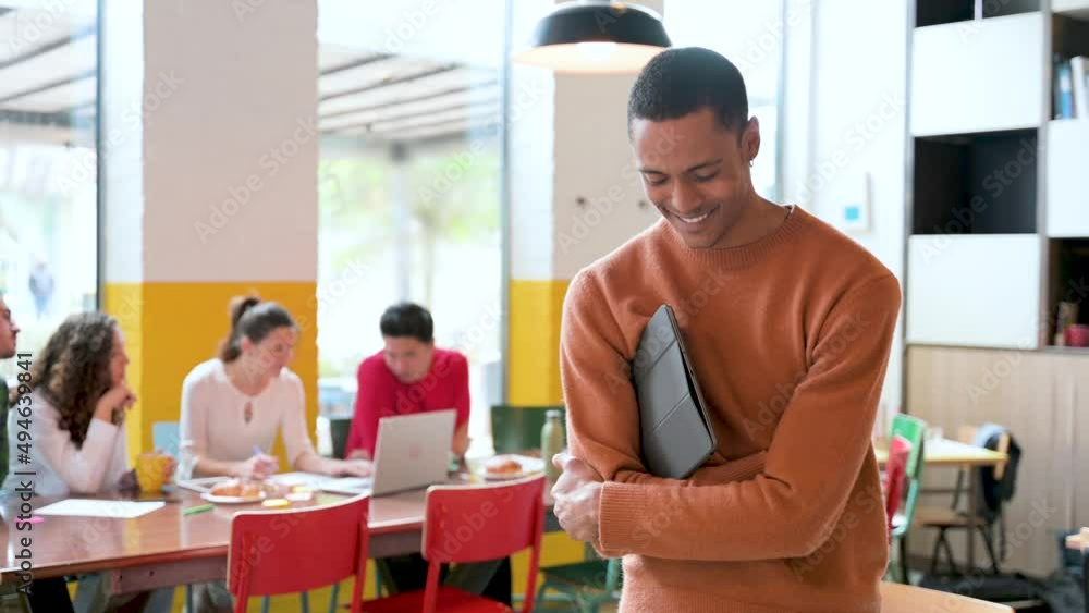 Portrait of Young African man in casual clothes keeping arms crossed holds tablet and looking at the camera with smile while group of people sitting at desk at background. Teamwork, co-working concept Portrait of Young African man in casual clothes keeping arms crossed holds tablet and looking at the camera with smile while group of people sitting at desk at background. Teamwork, co-working concept