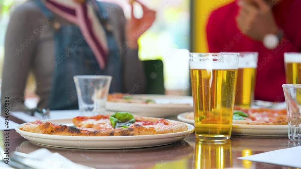 Close-up glass of bubbling beer and pizza on table with unrecognizable couple speaking at unfocused background. Close-up glass of bubbling beer and pizza on table with unrecognizable couple speaking at unfocused background.