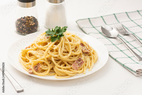 Cream and bacon Pasta with green parsley leaves on a white plate with a kitchen towel and salt and paper on background with fork, knife, and spoon. Italian spaghetti with pamezan.