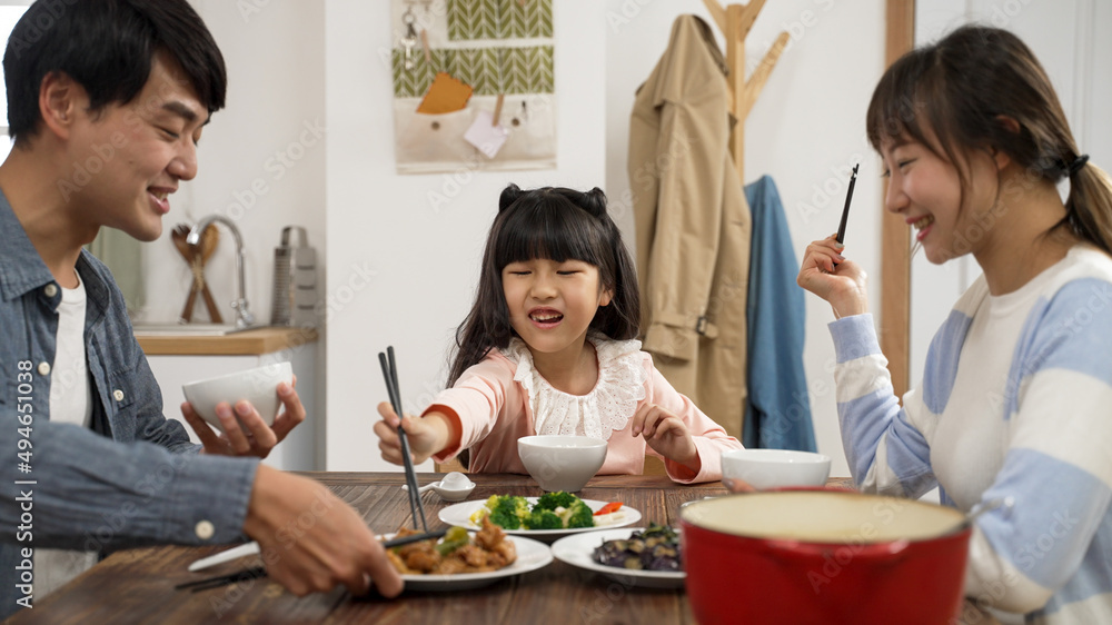 happy asian family of three talking and laughing while having lunch together at home. the dad helps pass the meat dish to her daughter and she accidentally drops the food happy asian family of three talking and laughing while having lunch together at home. the dad helps pass the meat dish to her daughter and she accidentally drops the food