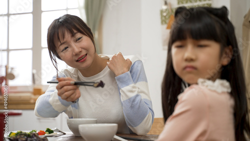 frustrated Asian mom trying to encourage daughter to eat vegetable in dining room at home. the girl turns head and refuses her worried mom. she don't like the vegetable
