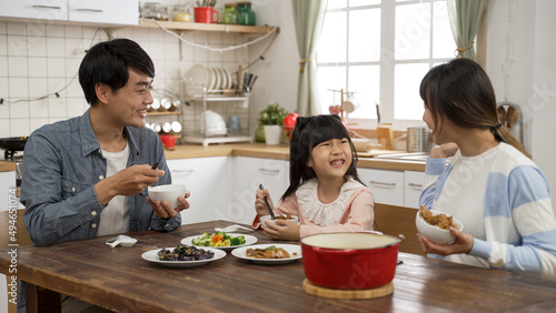 beautiful Asian family of three eating together in a modern dining room with daylight. caring father filling wife’s bowl with meat as the mother also fills daughter’s bowl with food