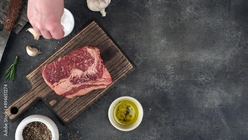 Chef sprinkling salt and pepper on ribeye steak, top view