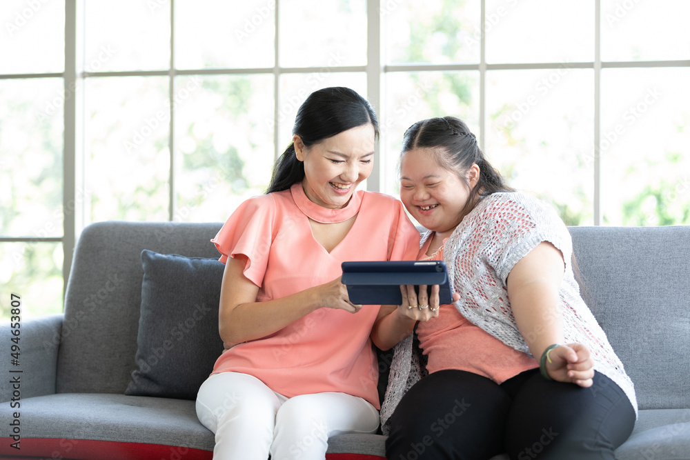 mother using tablet computer with a girl down syndrome or her daughter, smiling and enjoying on sofa mother using tablet computer with a girl down syndrome or her daughter, smiling and enjoying on sofa