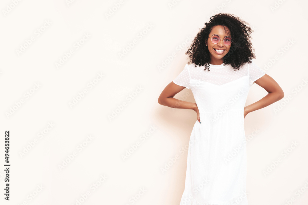 Beautiful black woman with afro curls hairstyle. Smiling model dressed in white summer dress. Sexy carefree female posing near wall in studio. Tanned and cheerful. Isolated