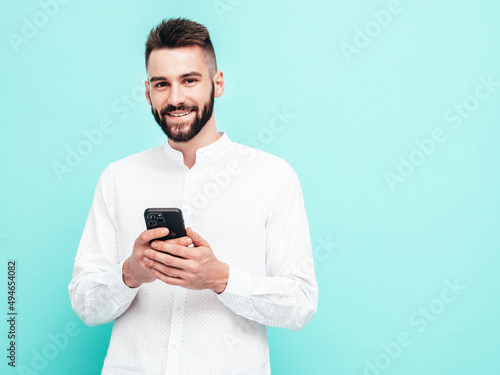 Handsome smiling model.Sexy stylish man dressed in shirt and jeans. Fashion hipster male posing near blue wall in studio. Holding smartphone. Looking at cellphone screen. Using apps