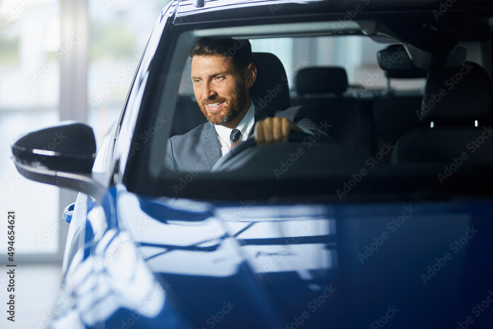 Positive bearded man in formal wear sitting on driver's seat of modern car and examining interior. Male customer choosing new transport at showroom.