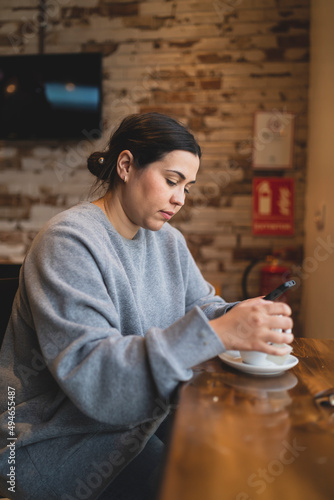 Chica joven guapa tomando té  en una cafetería muy acogedora con sudadera gris y mirando el smartphone