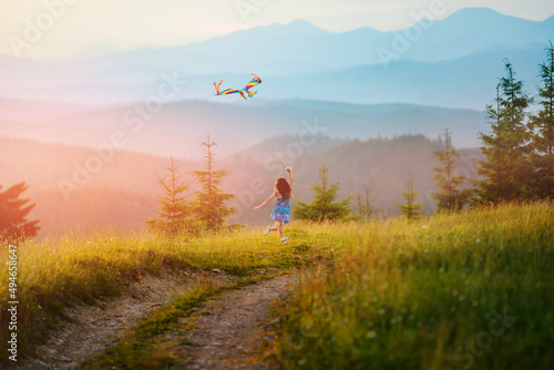Little happy child girl running with kite. Wonderful mountain landscape in Alps at sunset. Blue ridges of mountains on horizon.
