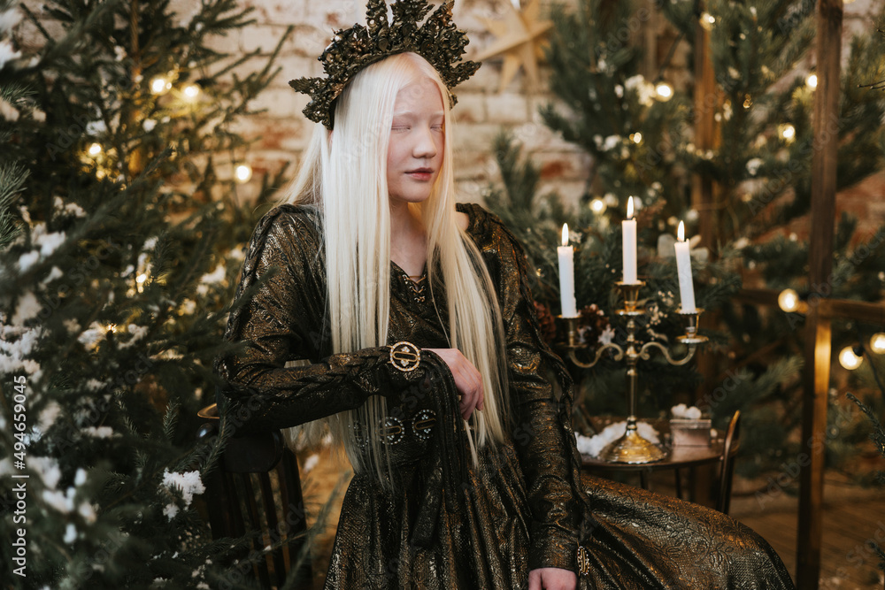 young albino woman with blue eyes and long white hair in beautiful green dress and crown stands in loft room decorated with wooden greenhouse and Christmas trees with twinkle lights, diverse people