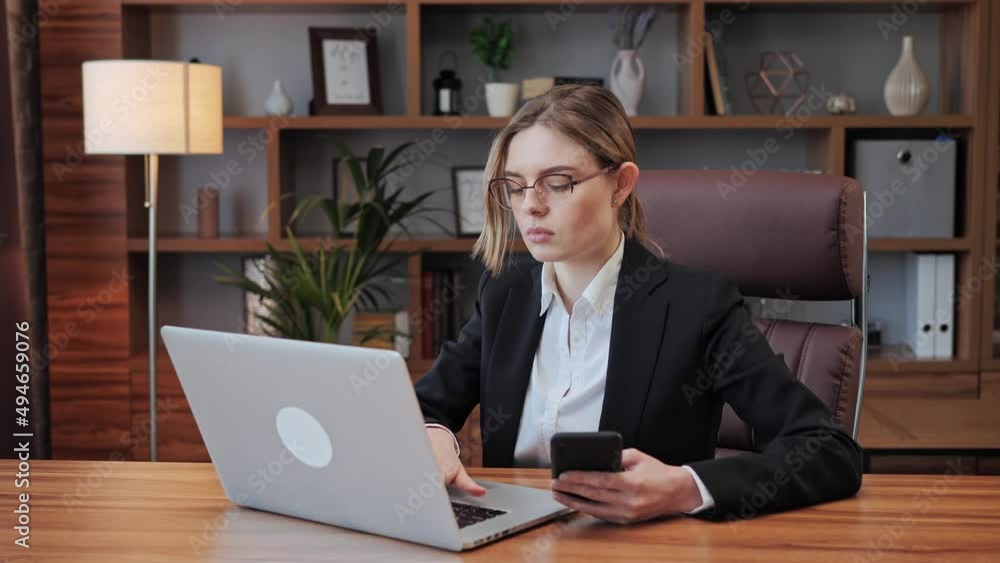 Smart business woman in glasses working from home distance online work. Girl using modern technology concept. Confident businesswoman working sits at desk look at laptop screen hold smartphone. Smart business woman in glasses working from home distance online work. Girl using modern technology concept. Confident businesswoman working sits at desk look at laptop screen hold smartphone.