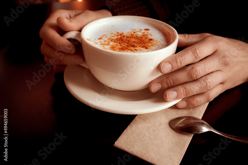 Female hands holding a cup of turkish traditional winter hot drink salep with cinnamon. White ceramic mug on plate and little spoon on napkin. Close-up. photo