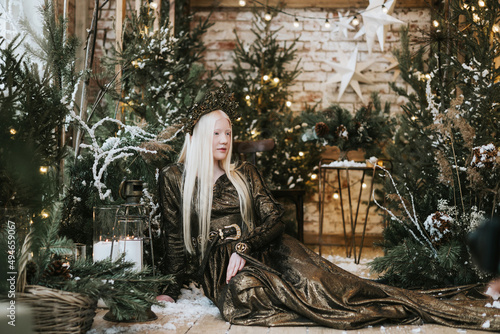 young albino woman with blue eyes and long white hair in beautiful green dress and crown stands in loft room decorated with wooden greenhouse and Christmas trees with twinkle lights  diverse people