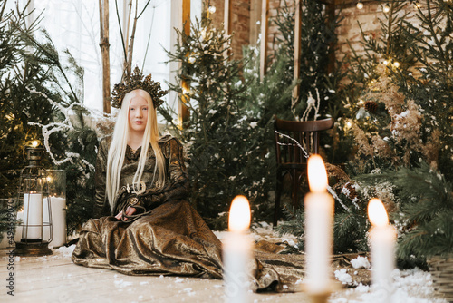 young albino woman with blue eyes and long white hair in beautiful green dress and crown stands in loft room decorated with wooden greenhouse and Christmas trees with twinkle lights, diverse people