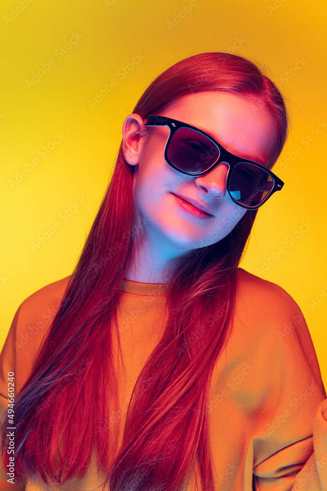 Portrait of trendy young girl posing in stylish black sunglasses isolated over yellow studio background