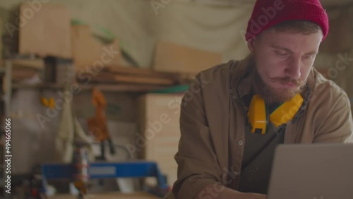 Young bearded carpenter with safety earmuffs over his neck using laptop in woodworking workshopYoung bearded carpenter with safety earmuffs over his neck using laptop in woodworking workshop photo