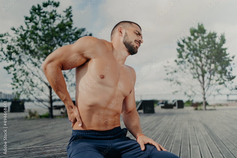 Obraz premium Sportsman touches lower back and feels backache after physical training exercises, poses outdoor