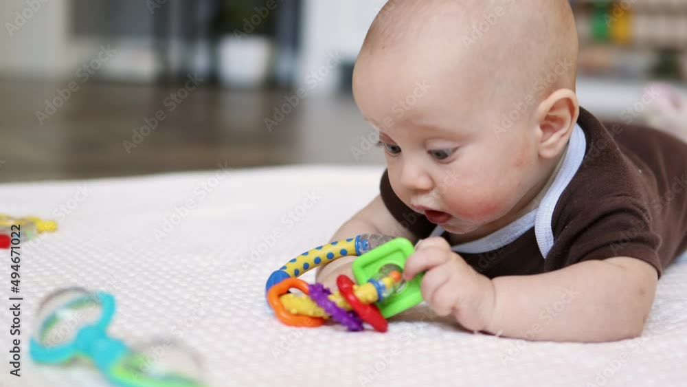 low angle view baby infant lying on tummy on white blanket indoors playing rattle toys. child trying reach colorful plastic noisemaker, learning to crawl, crying getting nervous, mad. kid's emotions low angle view baby infant lying on tummy on white blanket indoors playing rattle toys. child trying reach colorful plastic noisemaker, learning to crawl, crying getting nervous, mad. kid's emotions