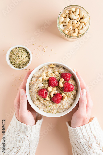 Bowl of oatmeal porridge with raspberries, hemp seeds and nuts in female hands over beige background. Top view. Concept of clean eating, dieting, healthy breakfast food