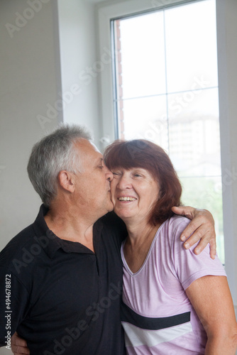 Elderly couple in casual clothes at light window of country house.