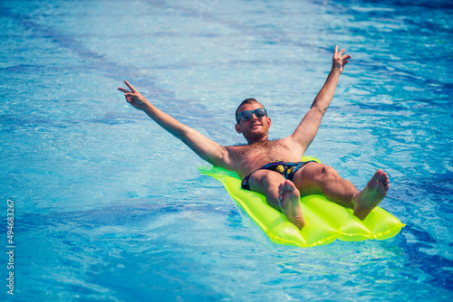 A young man bathes in the pool on a yellow inflatable mattress. Relax in the bright sun on vacation. Happy successful guy