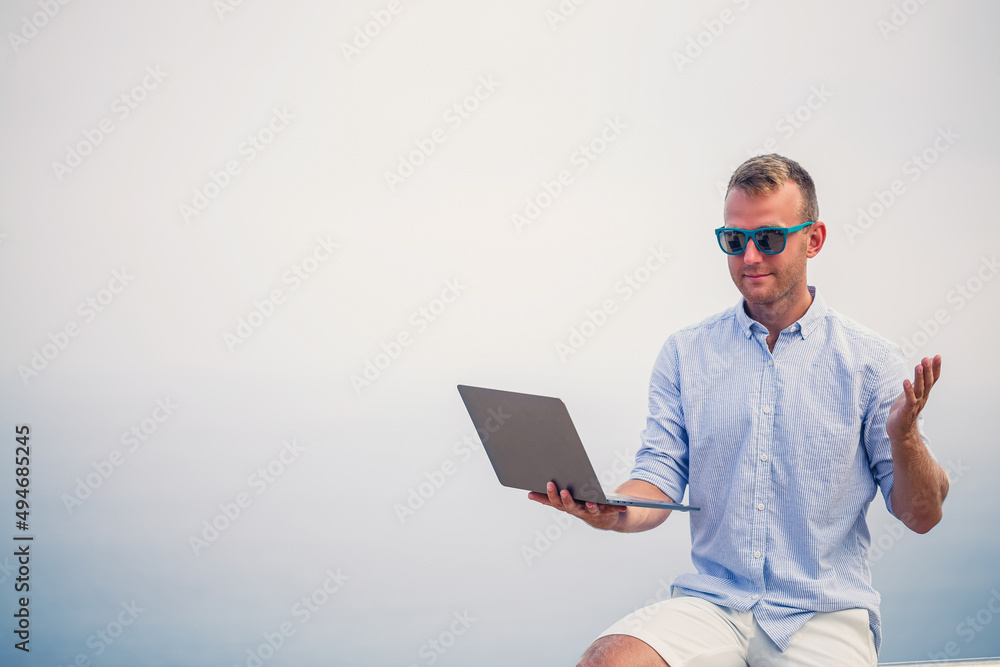 Young successful male businessman working with laptop on vacation by the sea. He wears sunglasses, a shirt and white shorts. Work outside the office, freelancer