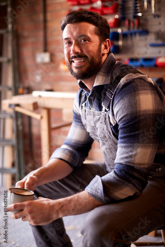 Portrait Of Smiling Man Wearing Overalls In Garage Workshop With Hot Drink